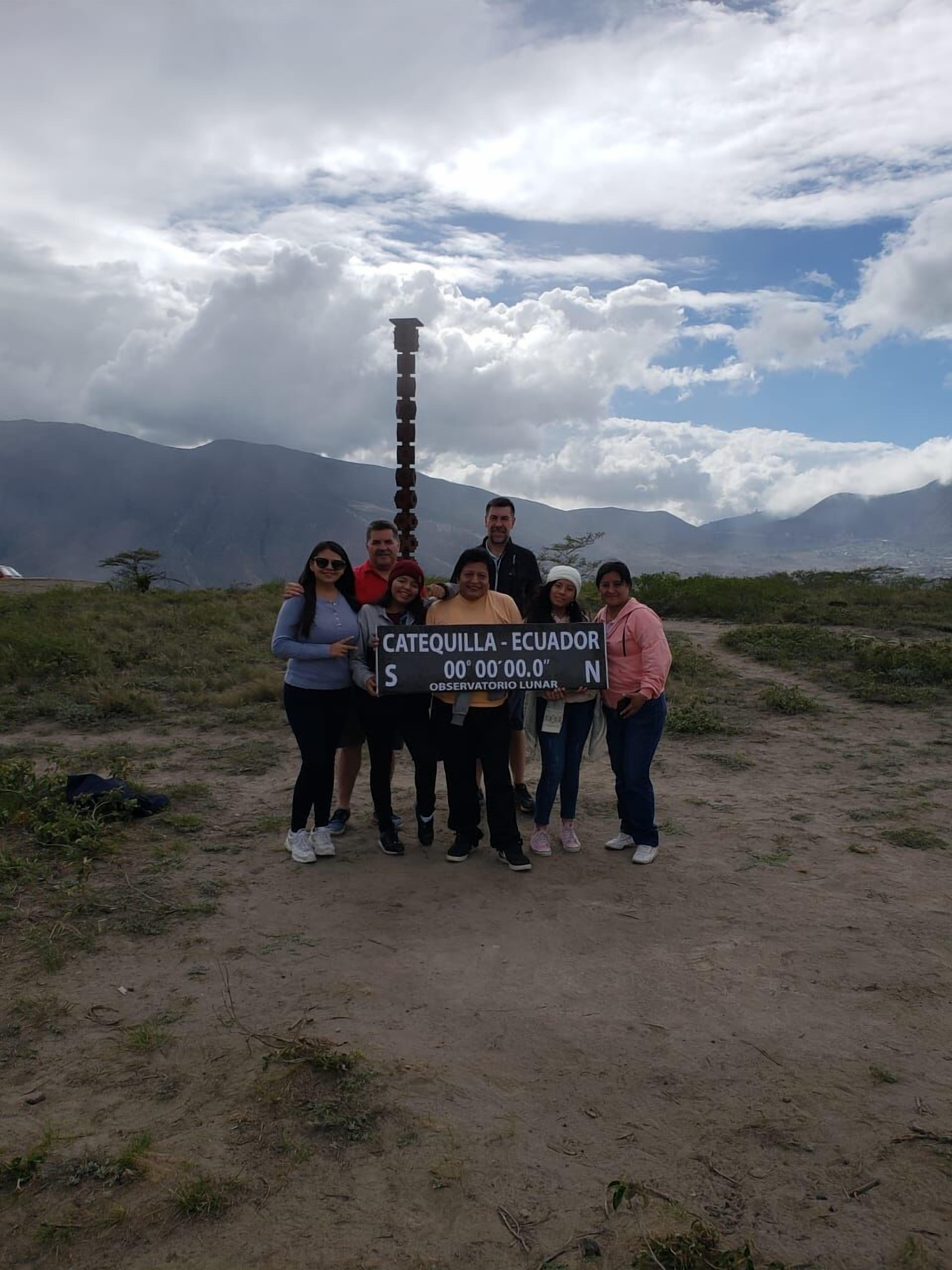 A group of people standing on a dirt path with a sign that reads "CATEQUILLA - ECUADOR 00°00'00.0" S OBSERVATORIO LUNAR."