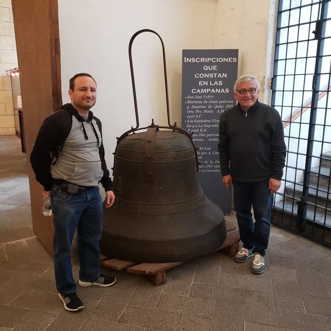 Two individuals standing beside a large bell with a sign listing inscriptions.