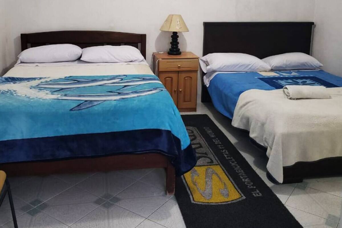 Habitación de hotel con dos camas, cada una con cubrecama azul y blanco. Hay una mesita de noche de madera con lámpara entre las camas. Un tapete negro y amarillo con el texto "EL PORTA CULLE" se ve en el suelo.