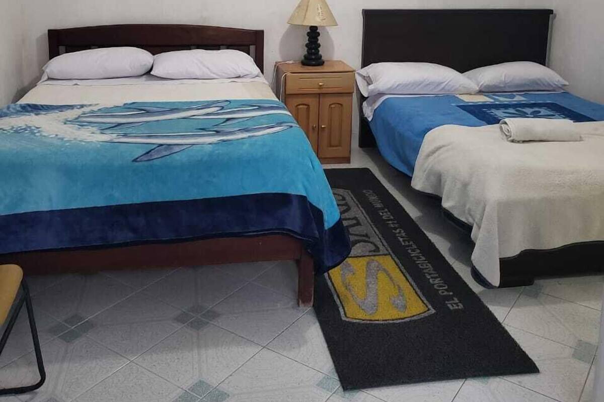 Una habitación con dos camas, cada una con una colcha azul y blanca. Hay una mesita con una lámpara entre las camas. El piso es de baldosas y el felpudo tiene un logotipo.