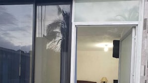 A room with a bed, a nightstand, a lamp, and a window with a view of palm trees. - Hostal Davinci Mitad del Mundo (San Antonio de Pichincha)