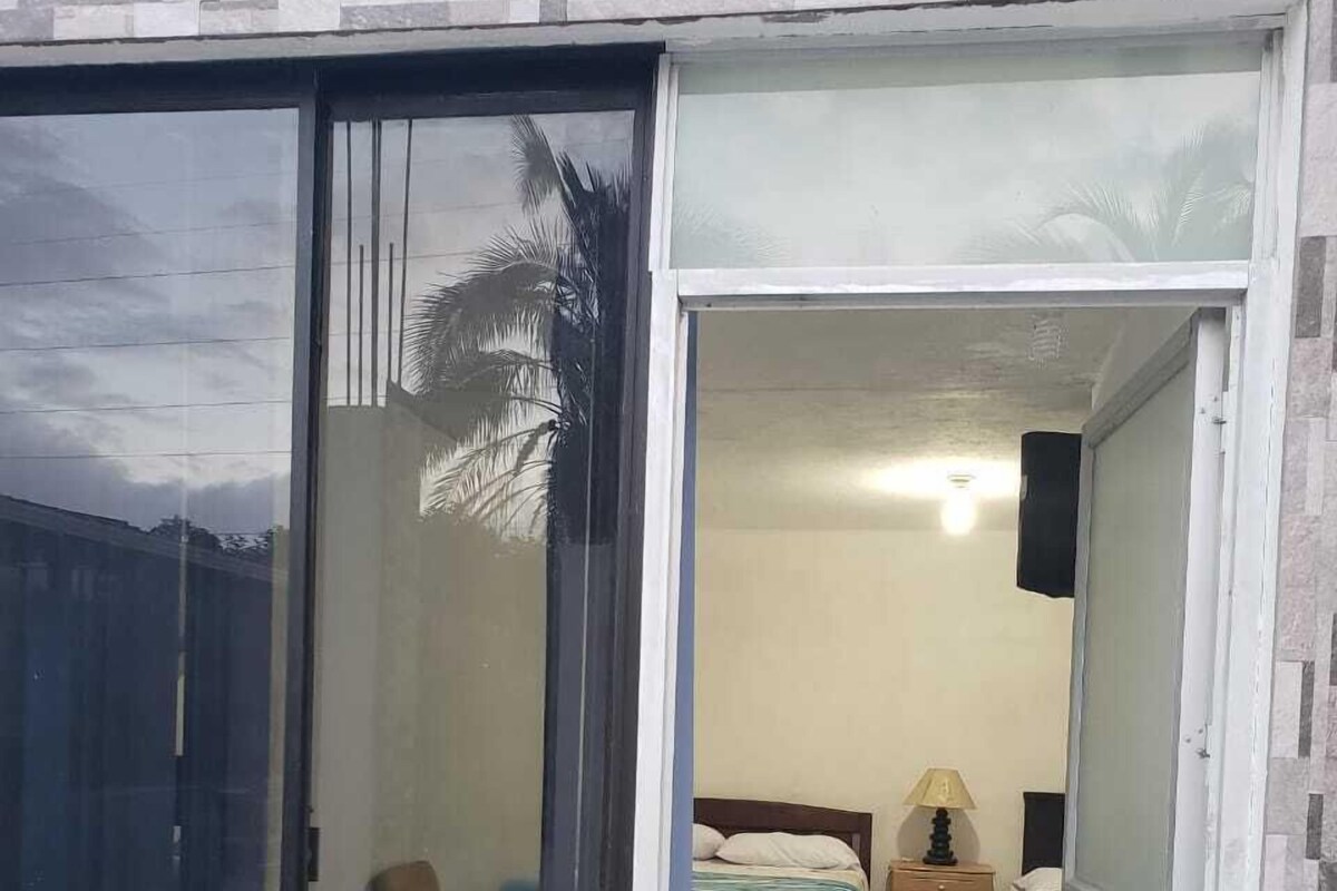 Quarto com cama, mesa de cabeceira, abajur e janela vista para palmeiras.