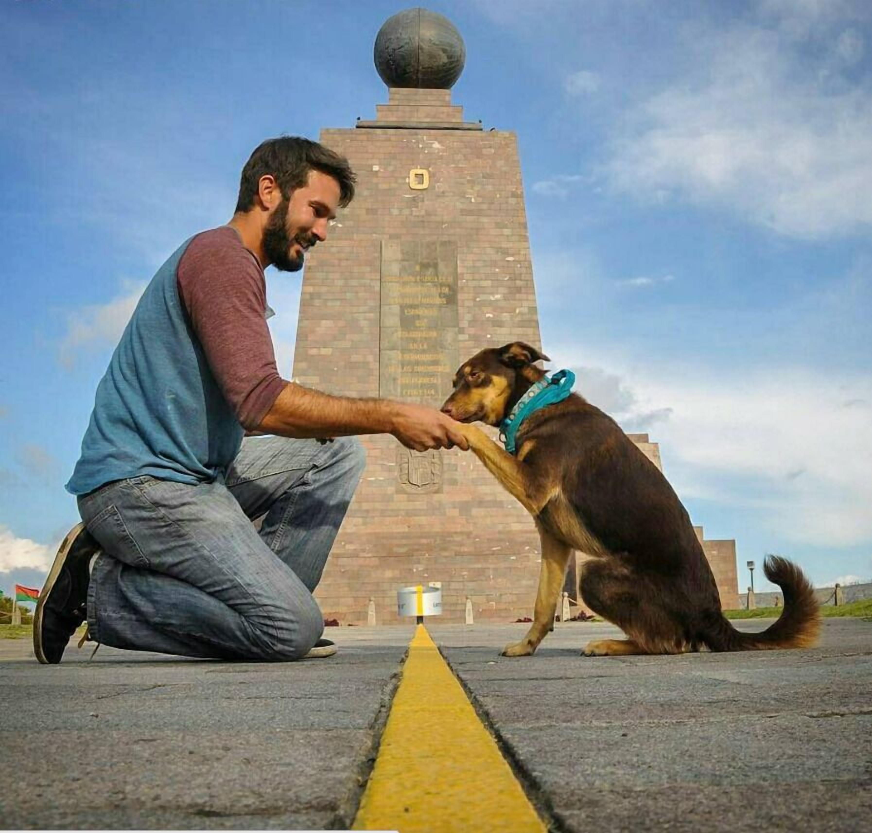 Un homme à genoux sur le sol, interagissant avec un chien devant un grand monument orné d’inscriptions.