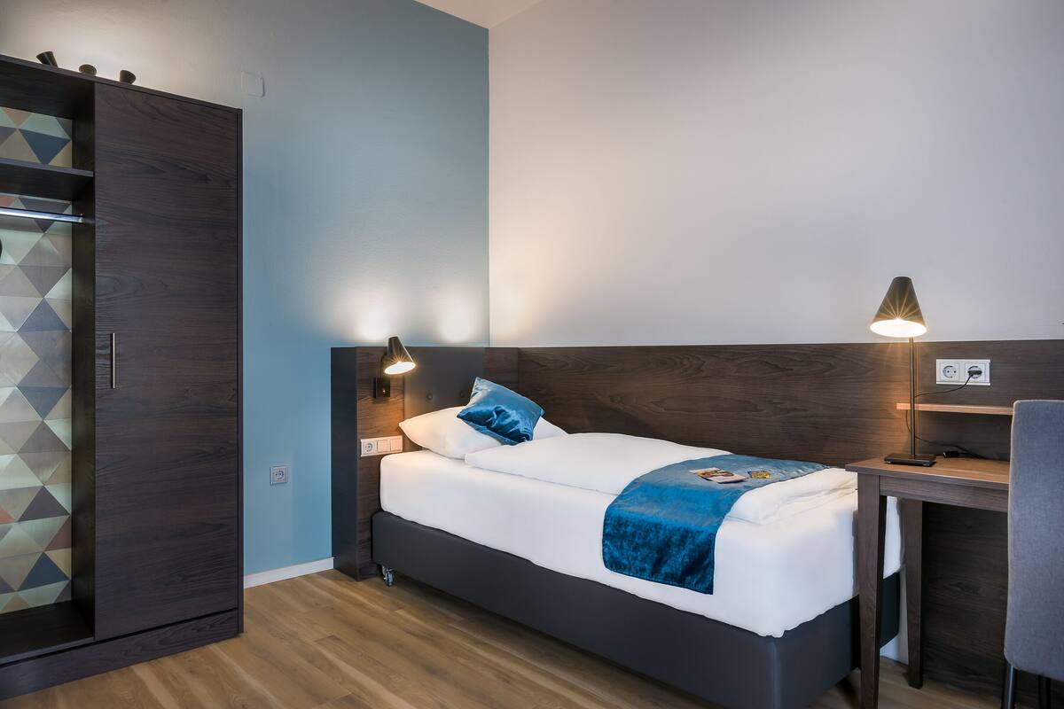 A modern hotel room with a bed, bedside tables, a desk, and a wardrobe.