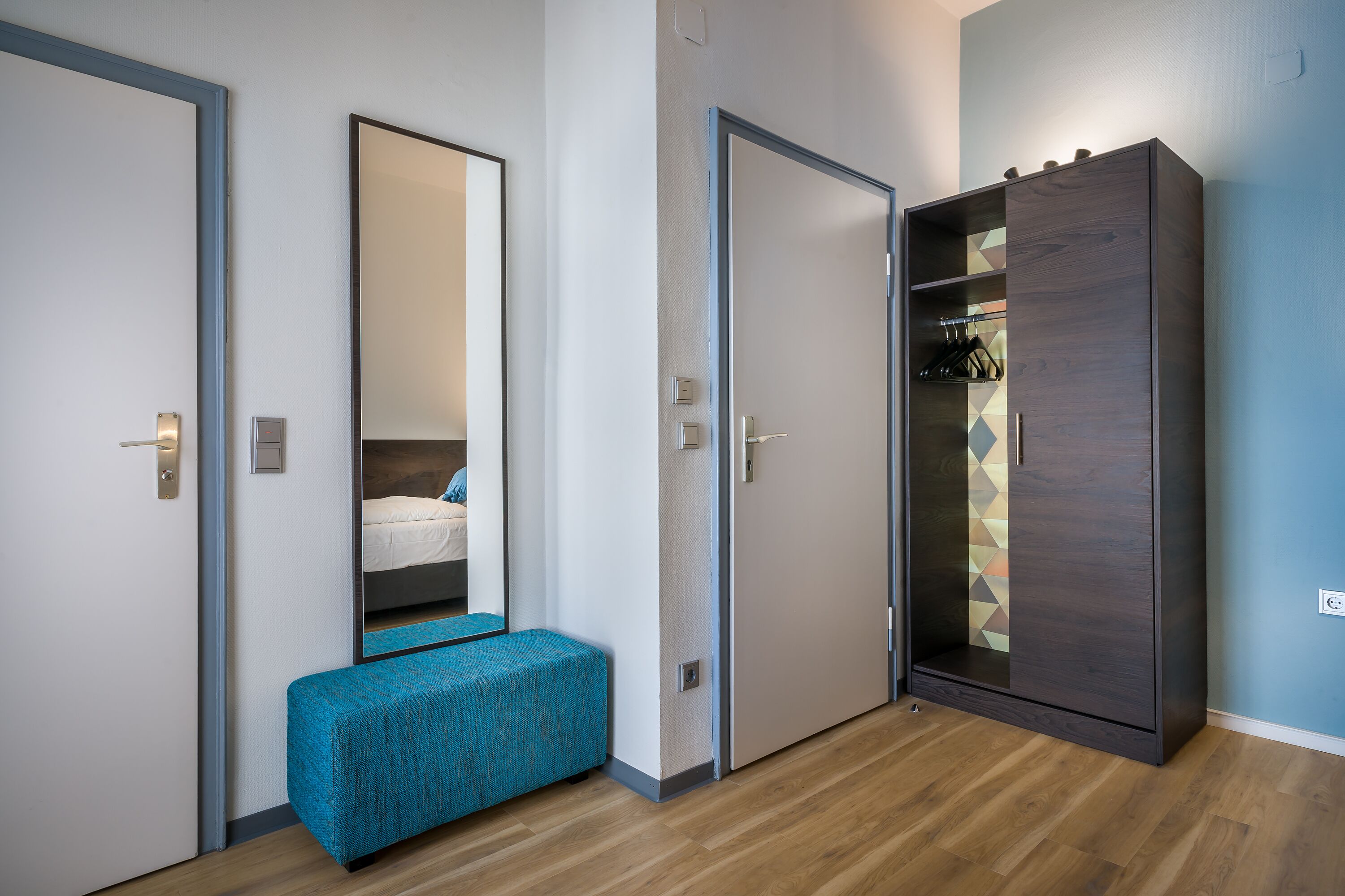 A hotel room with a wooden floor, a blue bench, a wardrobe, and a mirror reflecting a bed.