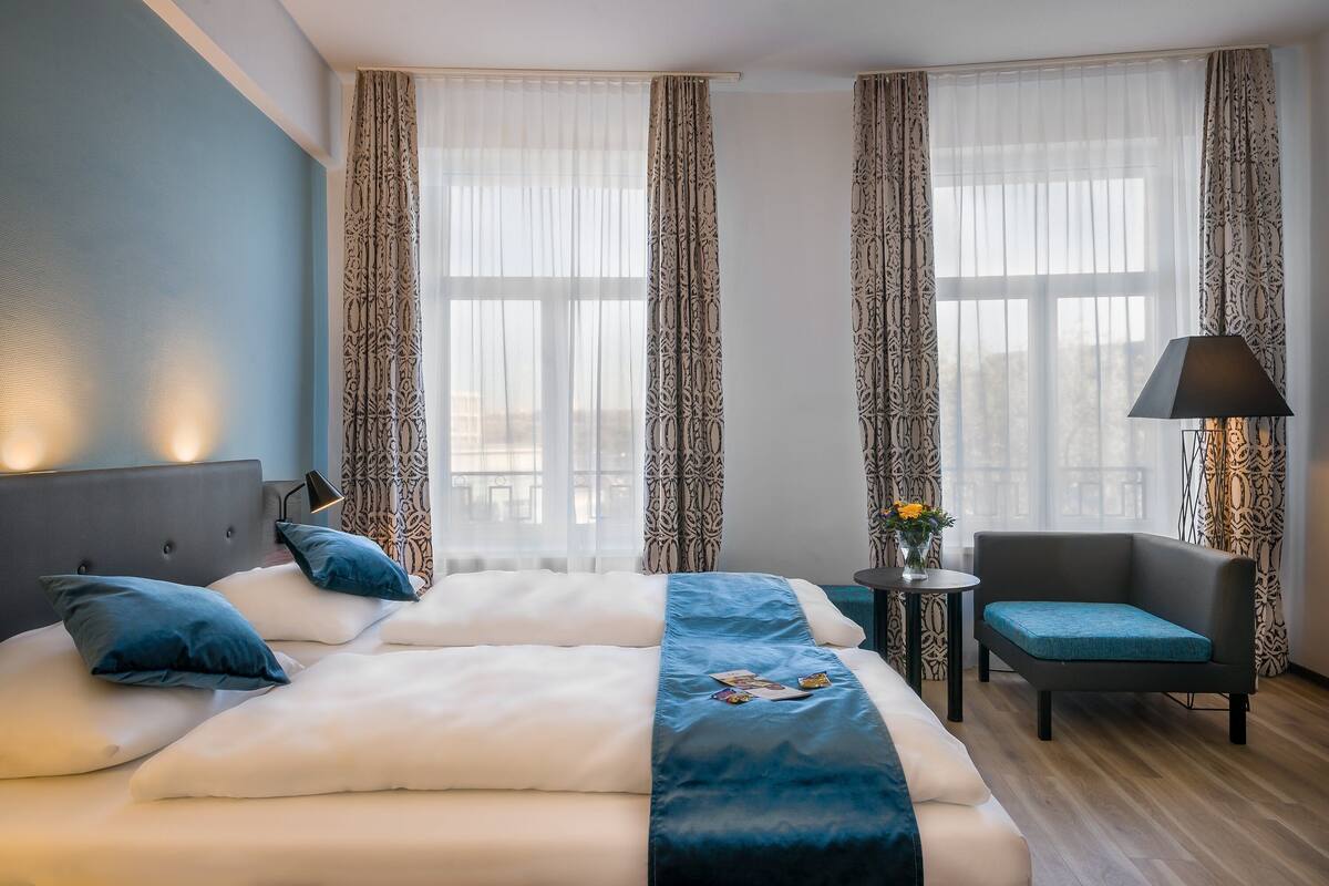 A hotel room with a large bed, a chair, a small table with a lamp, and patterned curtains.