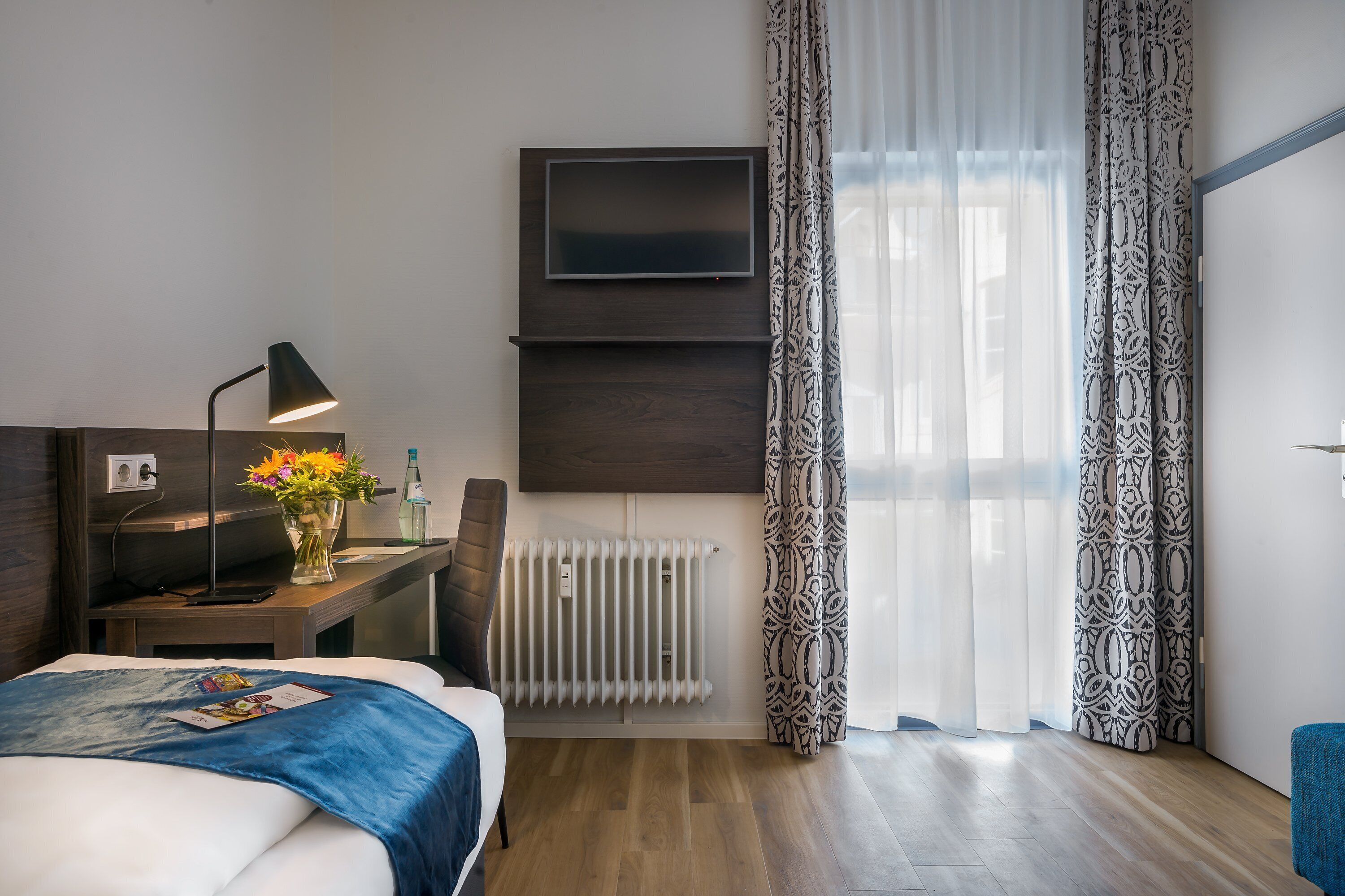 Ein modernes Hotelzimmer mit einem Bett, einem Schreibtisch mit Lampe, einem Flachbildfernseher, einer Heizung, gemusterten Vorhängen und einem Fenster mit Tageslicht.