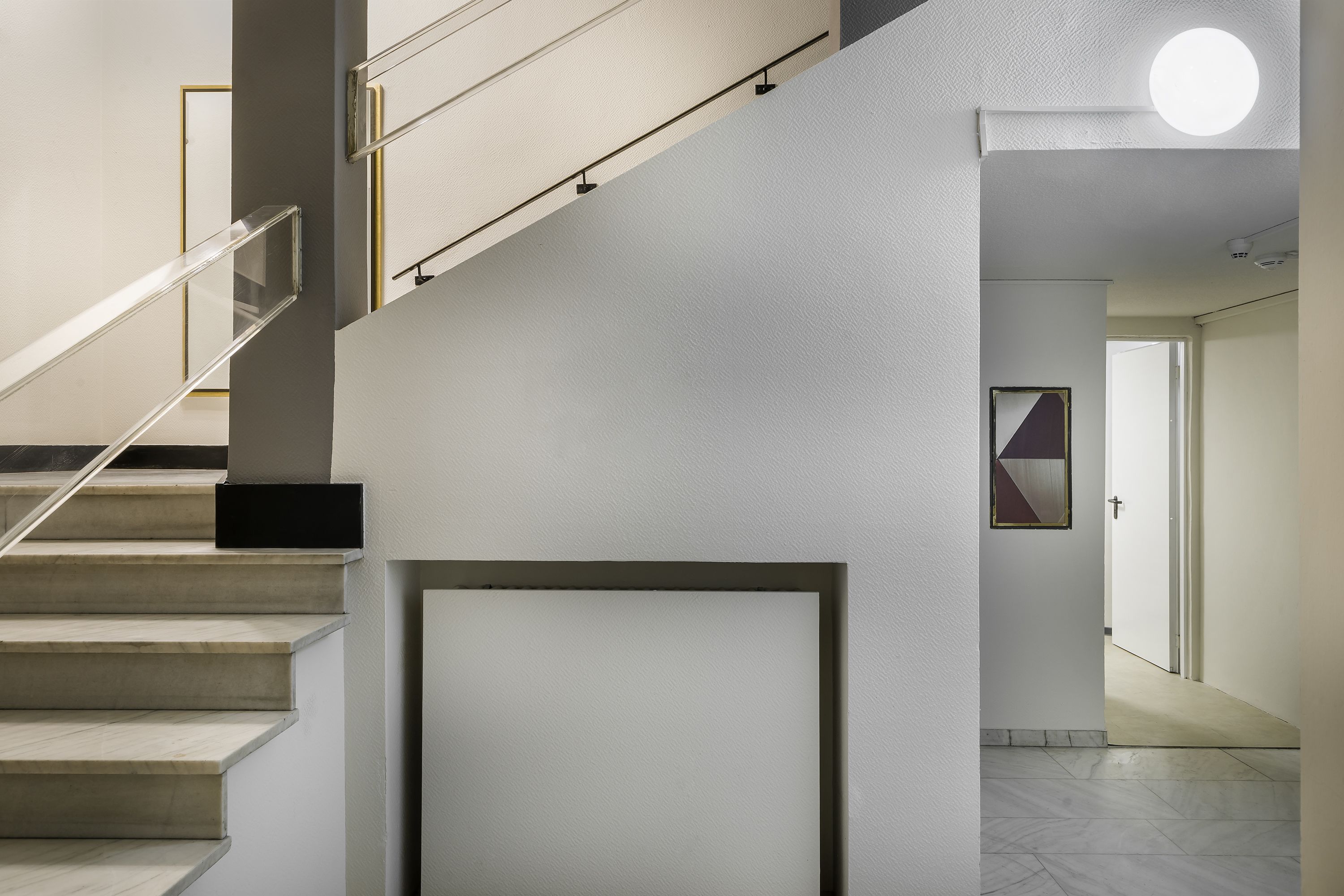 Una escalera moderna con barandilla de vidrio, un panel de pared geométrico y una lámpara de techo redonda.