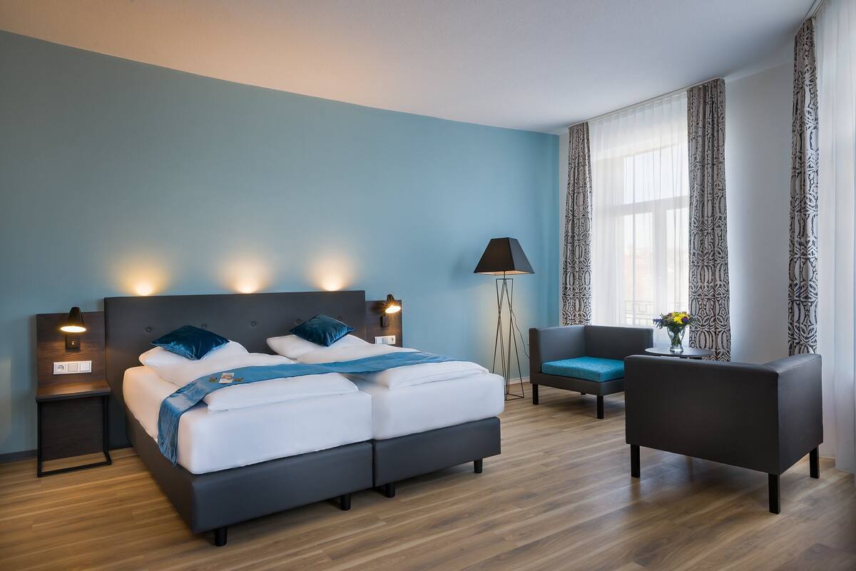 A modern hotel room with a large bed, a desk, a chair, and a lamp.