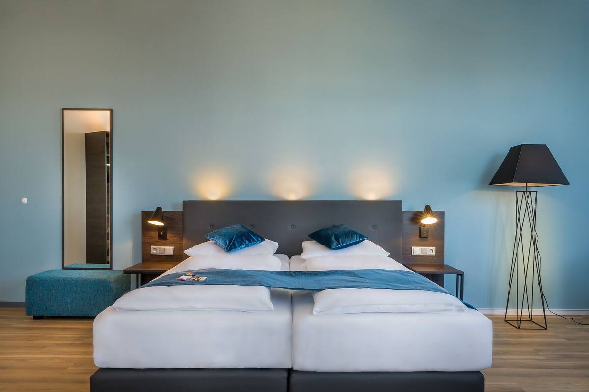 A modern hotel room with a large bed, two bedside tables with lamps, a floor lamp, and a large mirror on the wall.