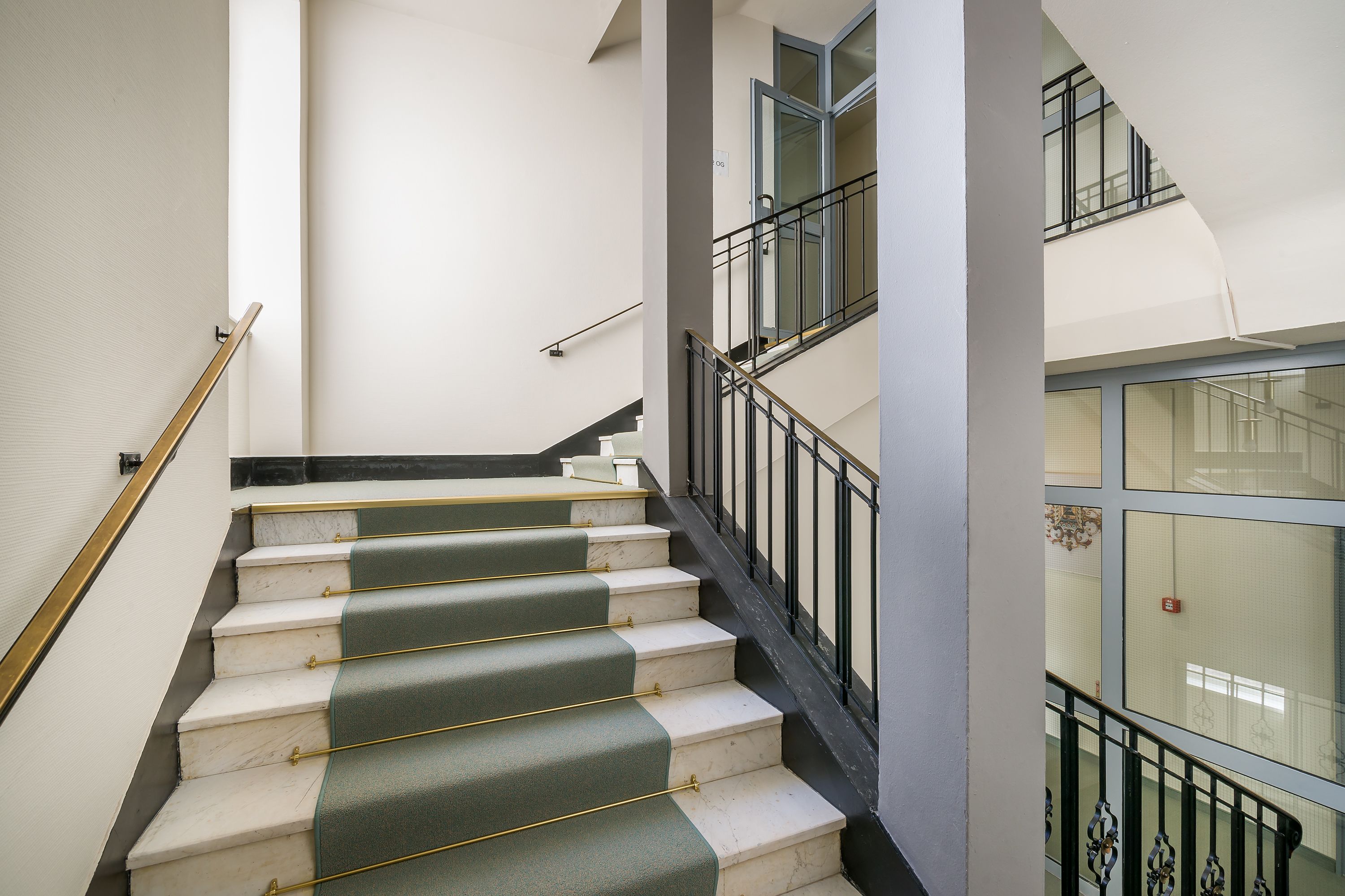 Una escalera de mármol con elegantes alfombras verdes, que conduce a un segundo piso con barandilla de vidrio.