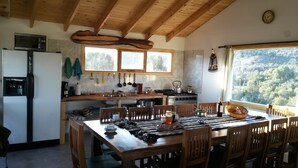 A rustic kitchen with wooden beams, a large window, and a dining area with wooden chairs. - Hostel Andino (Villa Pehuenia)