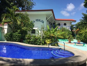 Una piscina con borde de azulejos, rodeada de abundante vegetación y un edificio con techo rojo y un cartel que dice "PERLITA".