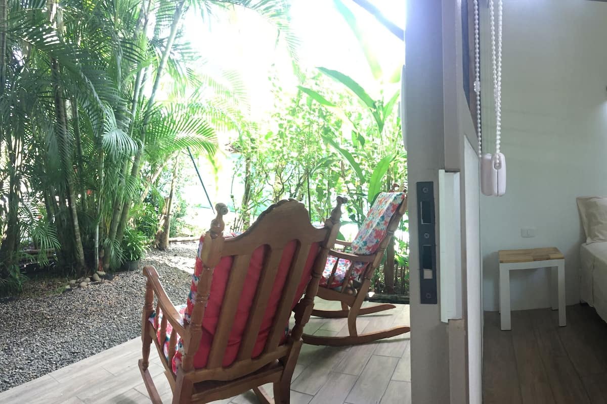 A bedroom with a bed, bedside tables, a small stool, a rocking chair, a refrigerator, and a window with a view of greenery.