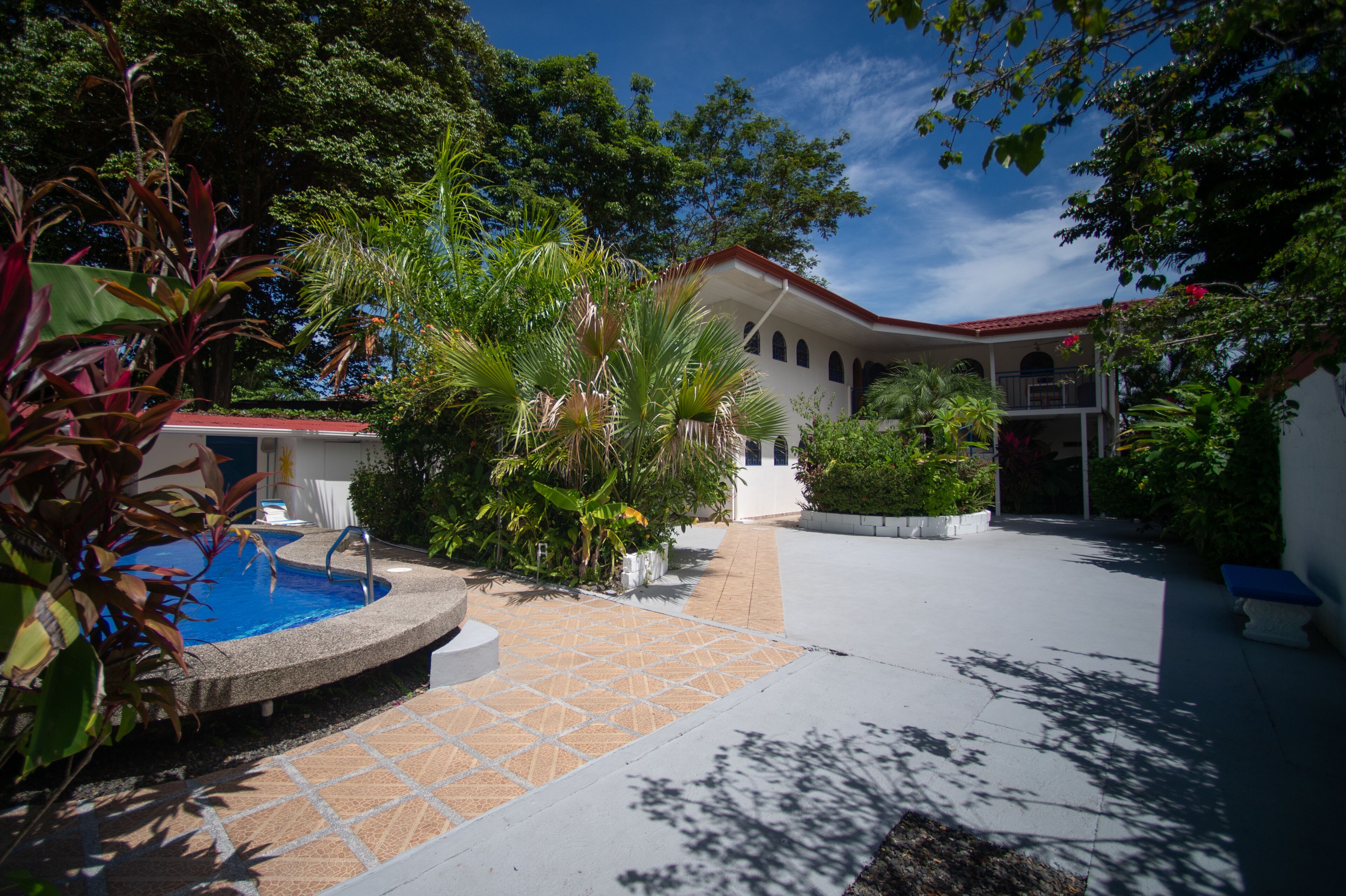 A pool area with a tiled deck, surrounded by lush greenery and a white building with a red roof.