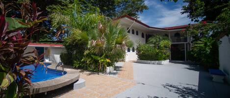 A pool area with a tiled deck, surrounded by lush greenery and a white building with a red roof.