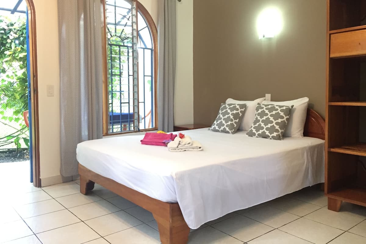 A bedroom with a bed, pillows, a nightstand, a window with curtains, and a view of greenery outside.