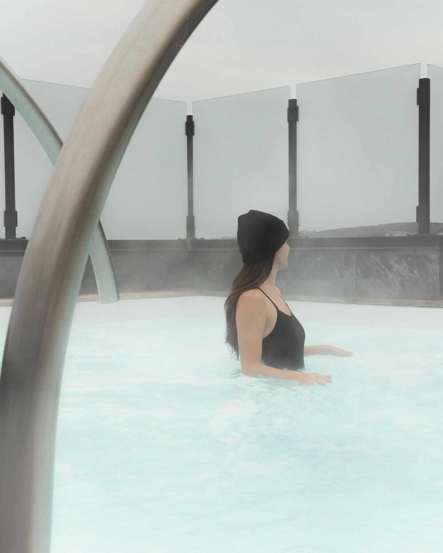 A person in a black swimsuit in a hot tub with steam, surrounded by a minimalist interior with columns and a plain wall.