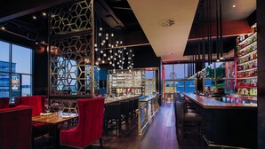 A modern bar with a wooden bar counter, red upholstered seating, and a view of the cityscape through large windows. - OTL Gouverneur Sherbrooke (Sherbrooke)