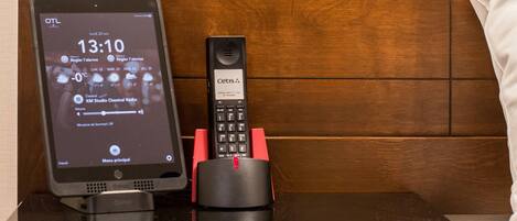 A hotel room with a wooden headboard, a bedside table with a tablet displaying the time and weather, a landline phone, and a remote control.