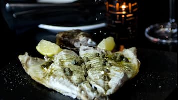Ein Teller Fisch mit Kapern und Zitronenscheiben, eine Kerze und ein Glas Wasser auf einem dunklen Tisch.