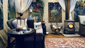 A cozy room with a wicker sofa, a small table with a candle, a decorative fan, and a wood-burning stove.