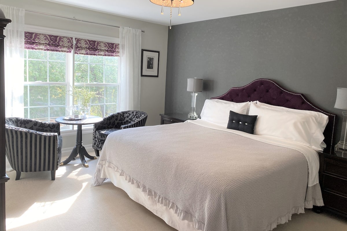 A bedroom with a large bed, a window with curtains, a chair, a table, and a lamp.