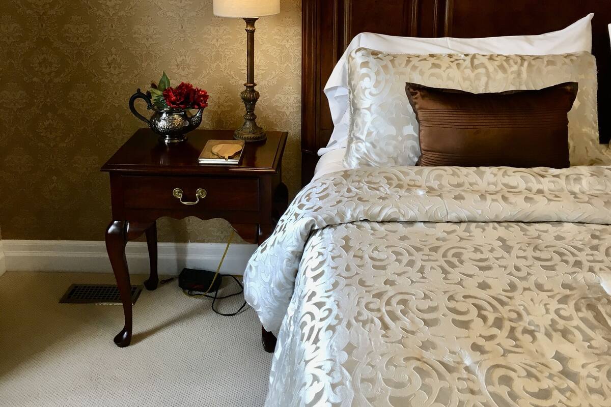 A hotel room with a wooden headboard, a bedside table with a lamp, a framed picture on the wall, and a floral-patterned bedspread.