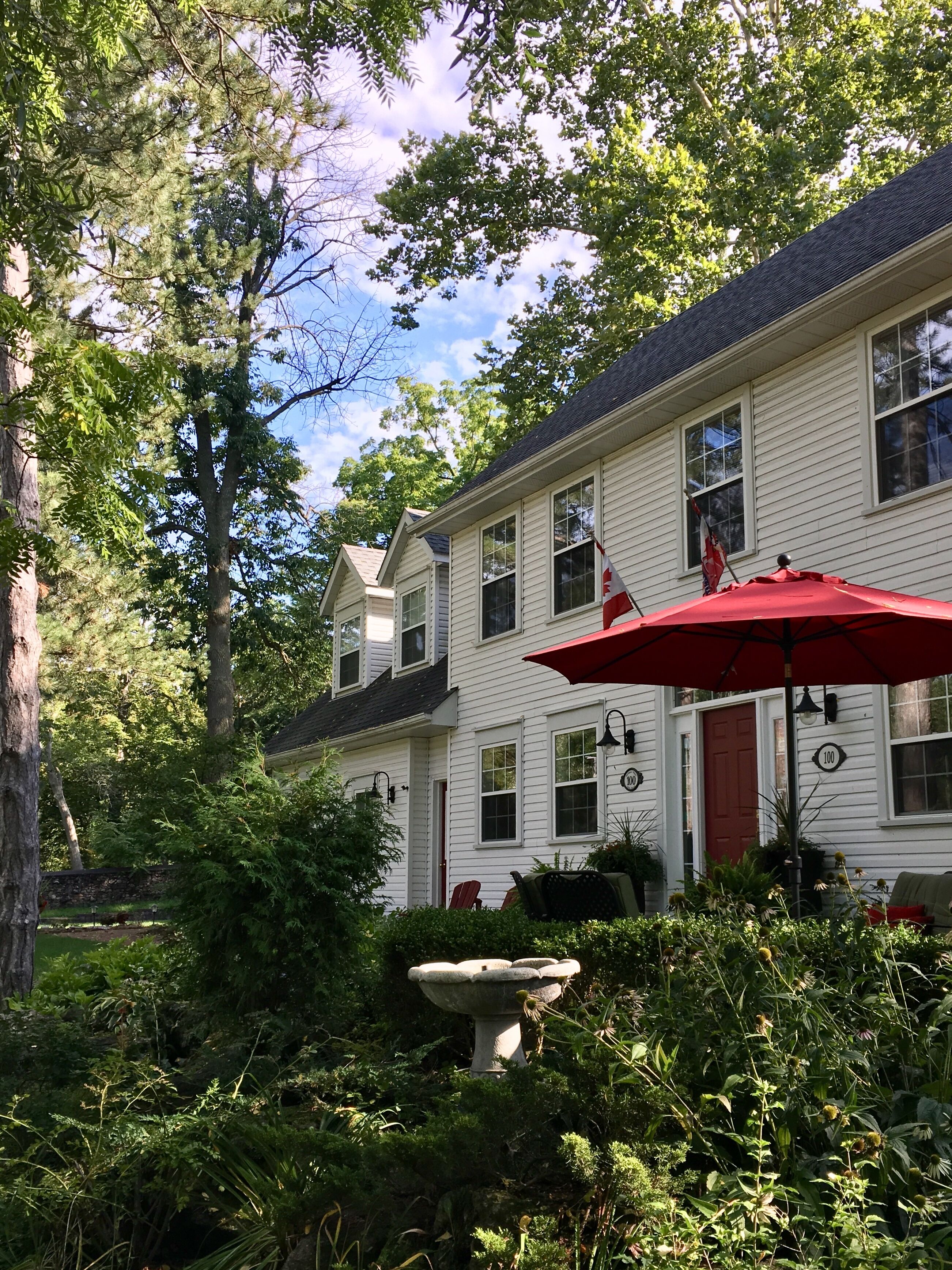 A white house with a red door, surrounded by lush greenery and a stone birdbath.