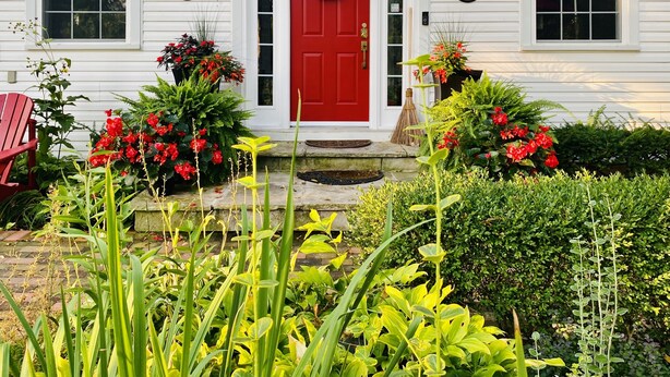 A white house with a red door, number 100, and a front porch with a red chair and potted plants.