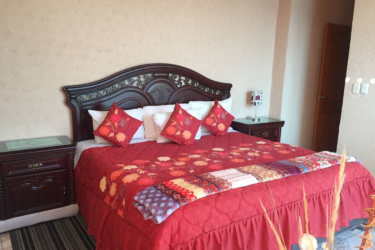 A neatly made bed with a red floral bedspread and decorative pillows, a wooden headboard, and a nightstand with a lamp.