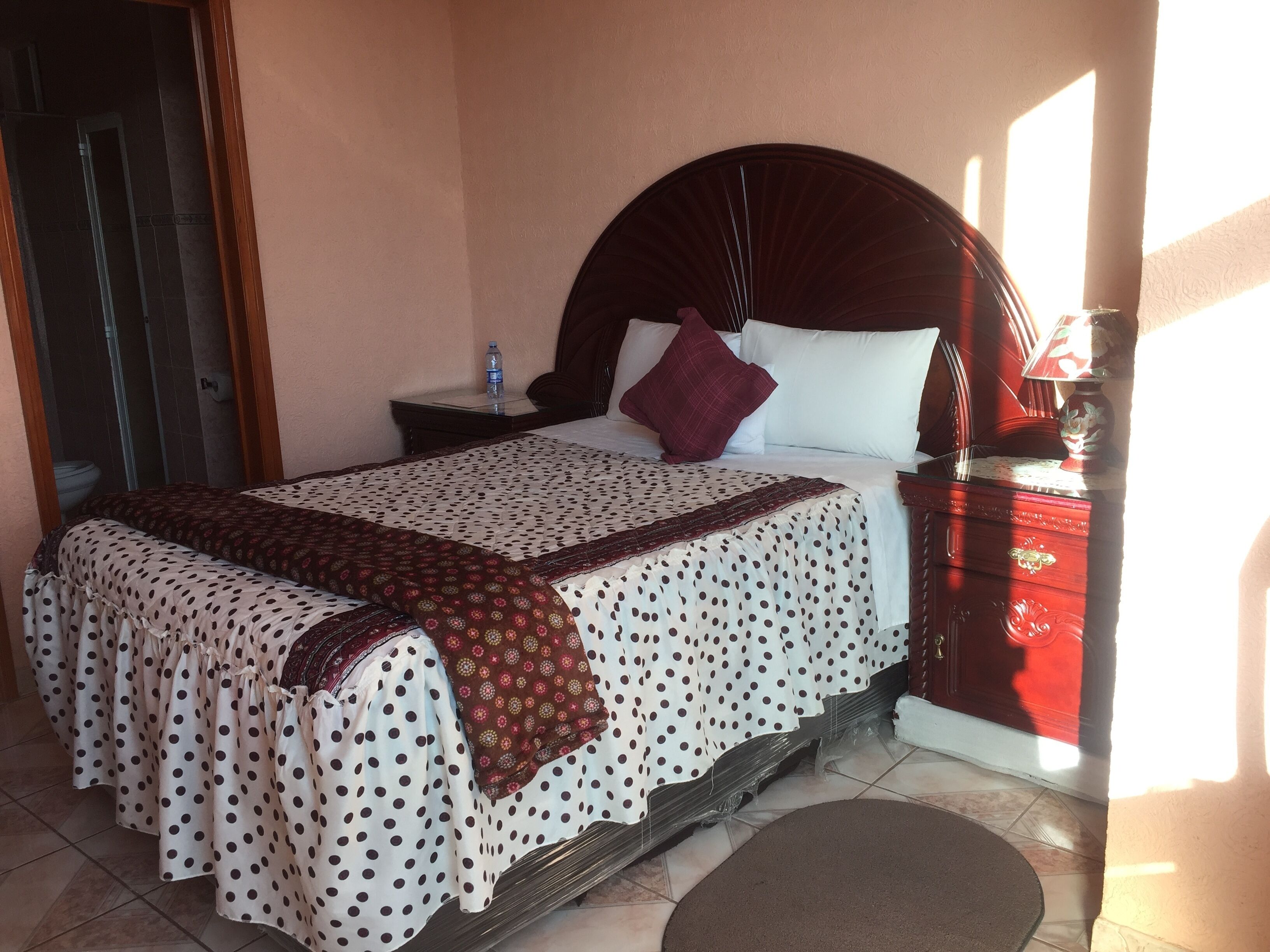 A neatly made bed with a polka dot bedspread and a brown throw blanket, a bedside table with a water bottle, and a nightstand with a lamp.