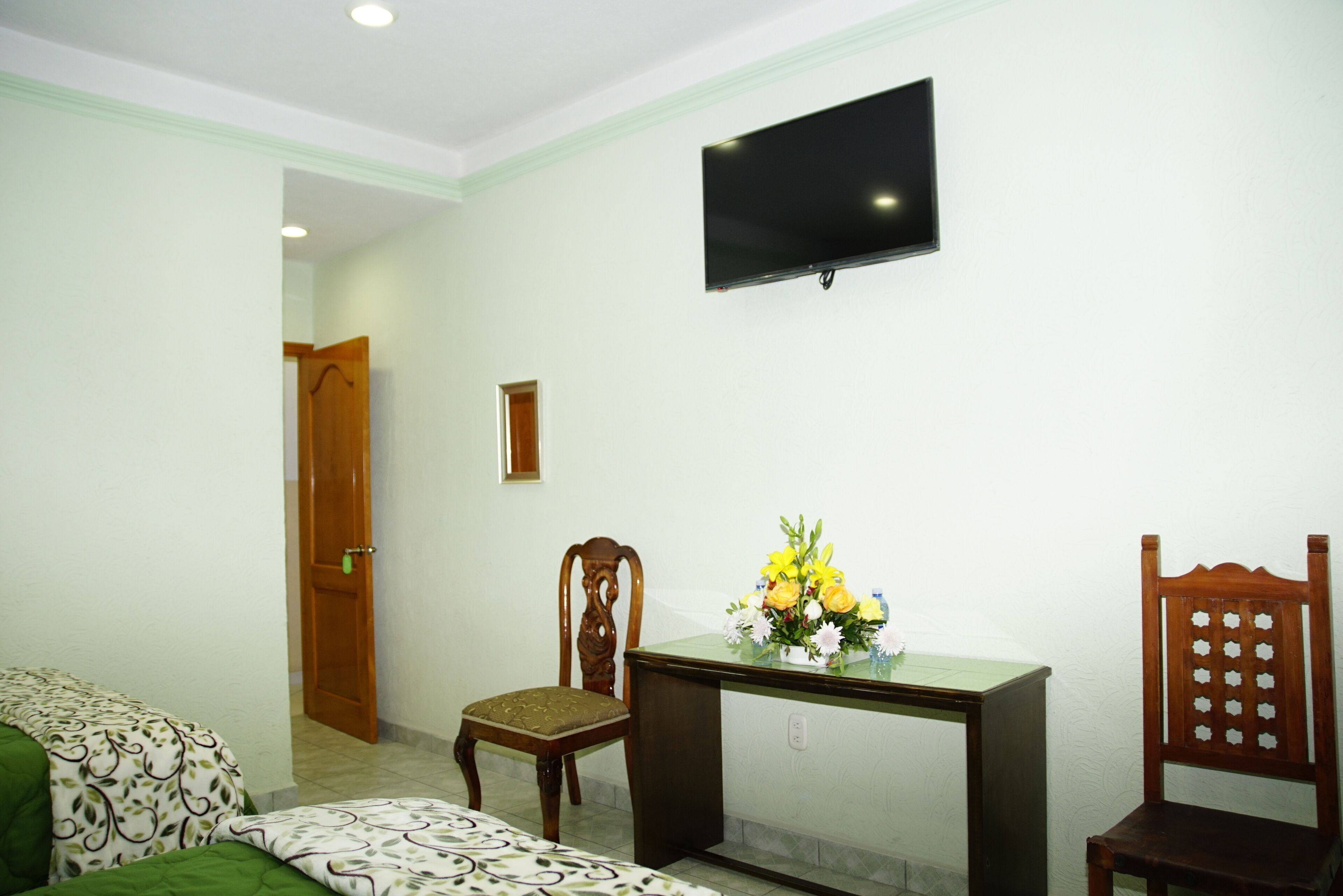 Habitación de hotel con cama, escritorio con un florero, televisor en la pared y puerta de madera.