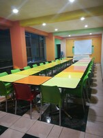 Sala de conferências com várias mesas e cadeiras, uma tela de projeção e um bebedouro.