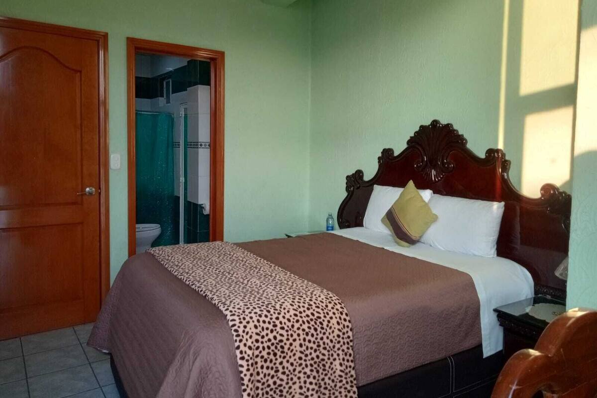 A bedroom with a wooden bed, a green wall, a bathroom visible through an open door, and a tiled floor.