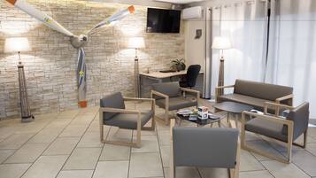 A modern hotel room with a stone wall, a desk, two chairs, a sofa, and a TV mounted on the wall.
