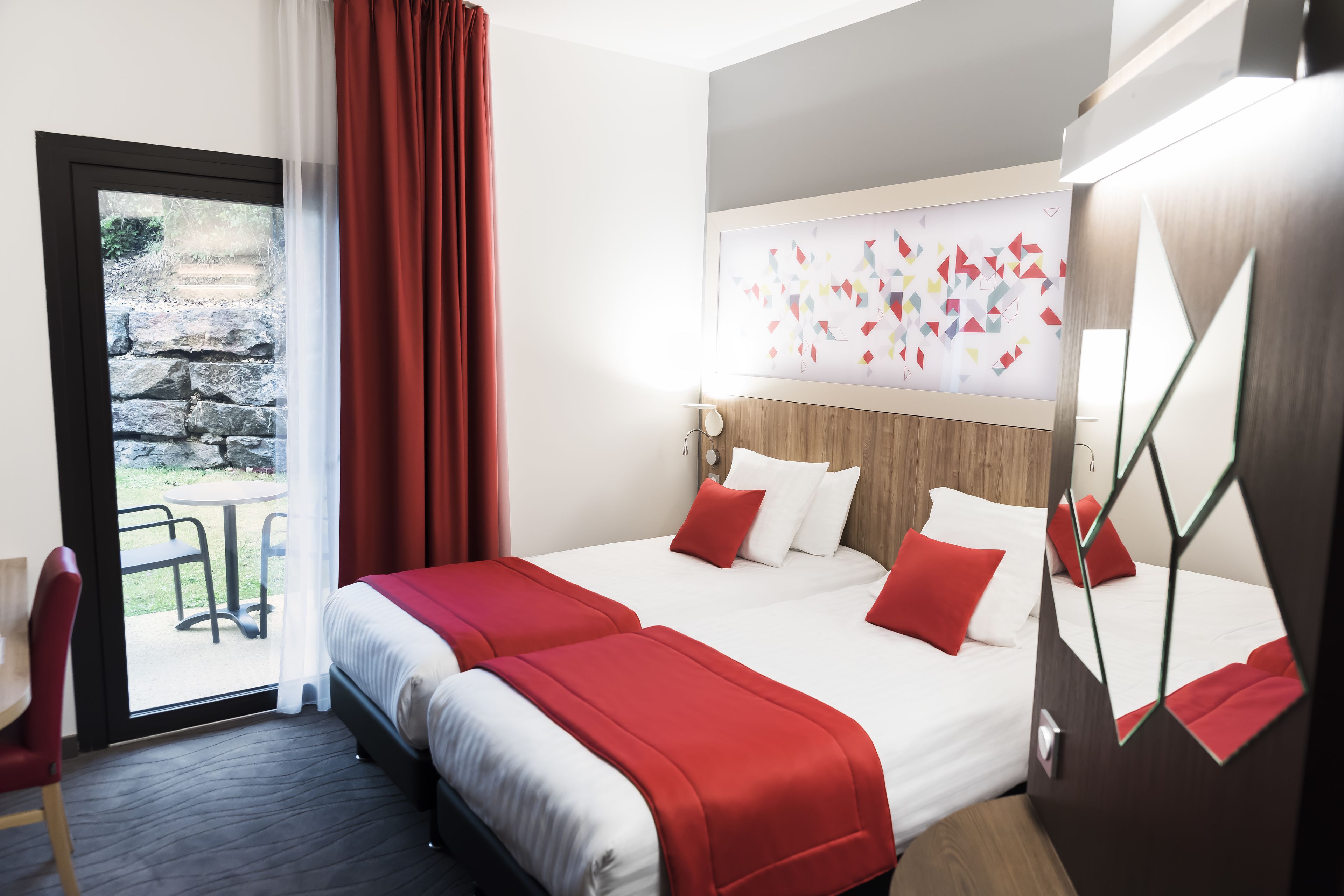 Hotellihuone, jossa on kaksi sänkyä, punaiset vuodevaatteet, suuri peili ja näkymä ulkona olevaan kiviseen portaikkoon.