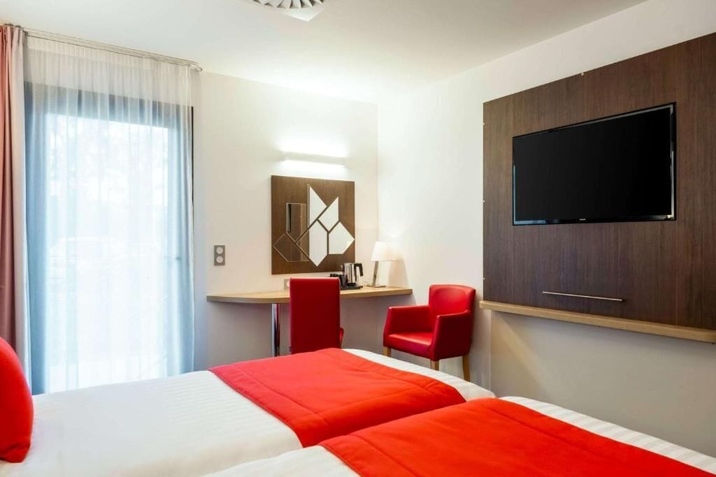 Een hotelkamer met een bed, een bureau, twee rode stoelen, een televisie en een raam met gordijnen.