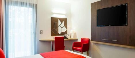 A hotel room with a bed, a desk, two red chairs, a TV, and a window with curtains.