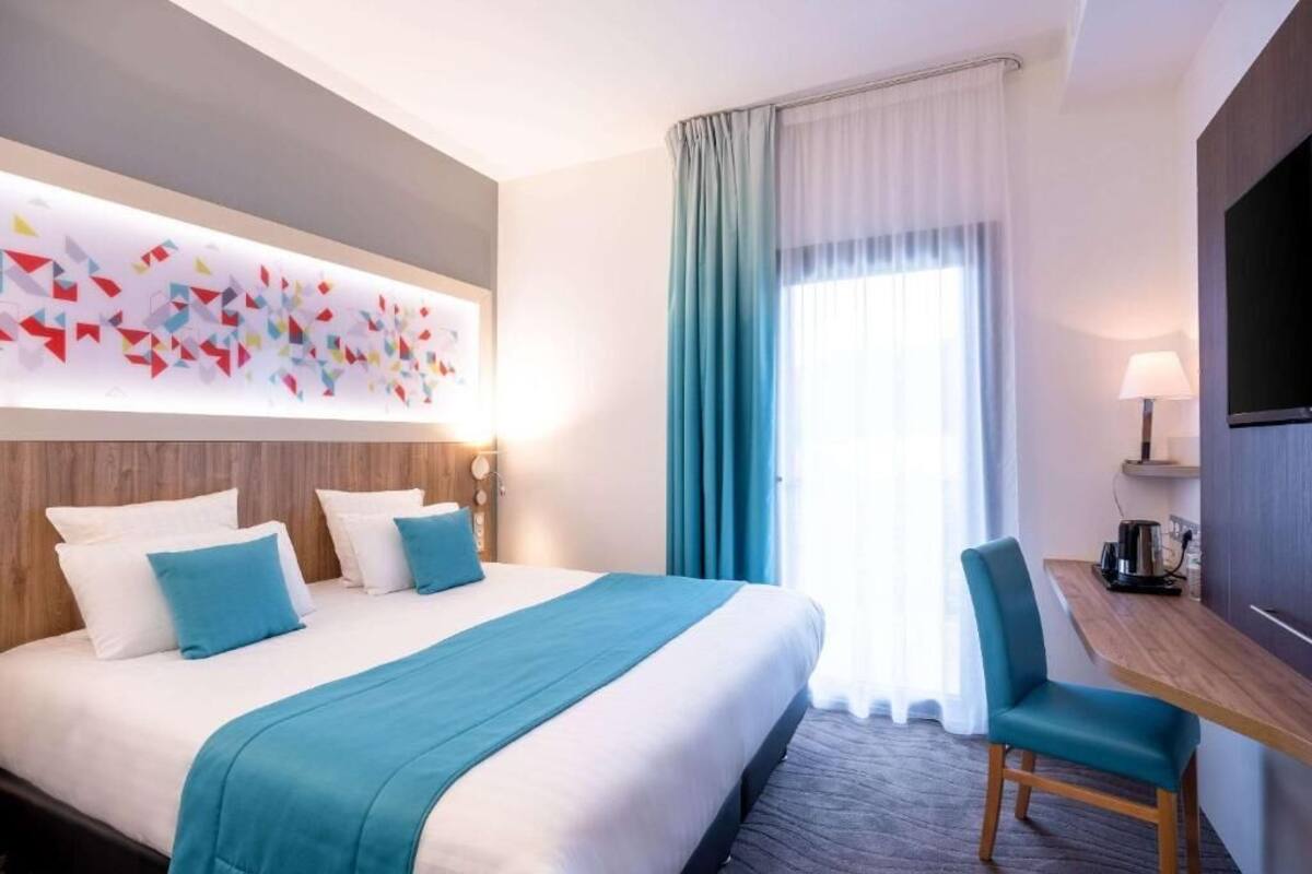 A hotel room with a large bed, a desk, a chair, a TV, and a wall art piece.