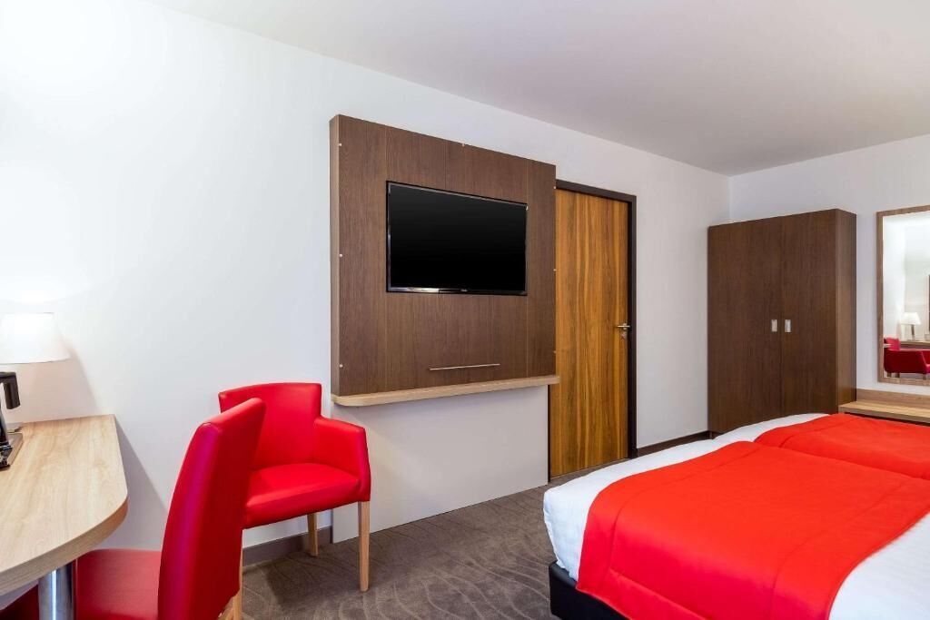 A hotel room with a bed, a TV mounted on a wall, a red chair, and a wooden desk with a lamp.