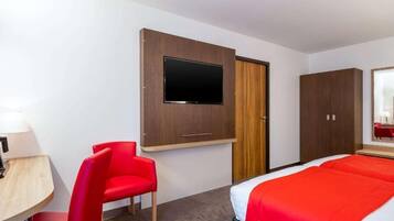 Une chambre d’hôtel avec un lit, un téléviseur fixé au mur, une chaise rouge et un bureau en bois avec une lampe.