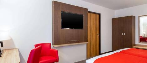 A hotel room with a bed, a TV mounted on a wall, a red chair, and a wooden desk with a lamp.