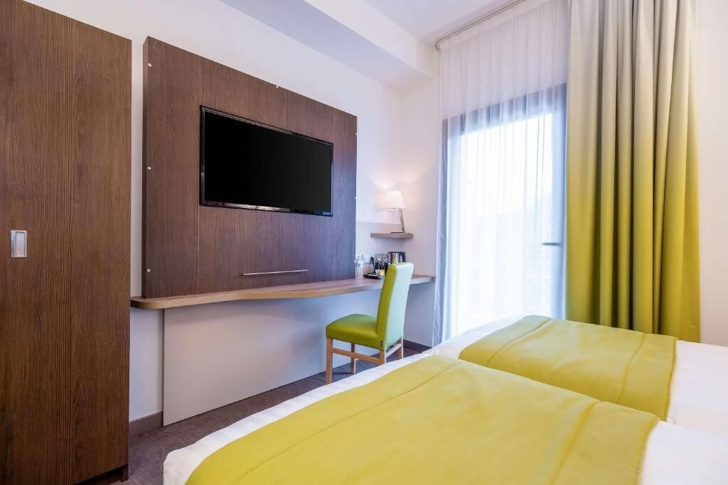 Habitación de hotel con cama, escritorio, silla, televisión y ventana con cortinas.