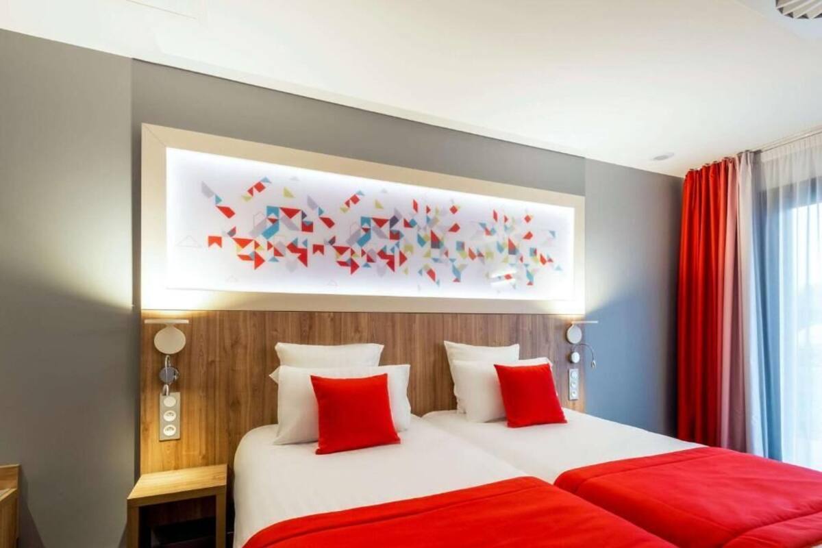A hotel room with a large bed, two red pillows, a wooden headboard, and a wall-mounted abstract art piece.
