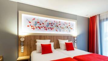 A hotel room with a large bed, two red pillows, a wooden headboard, and a wall-mounted abstract art piece.