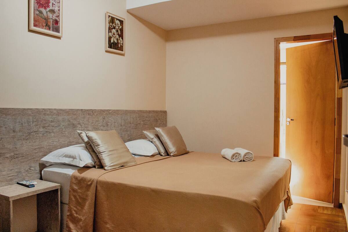 A neatly made bed with a headboard, a bedside table with a remote control, and two framed pictures on the wall.