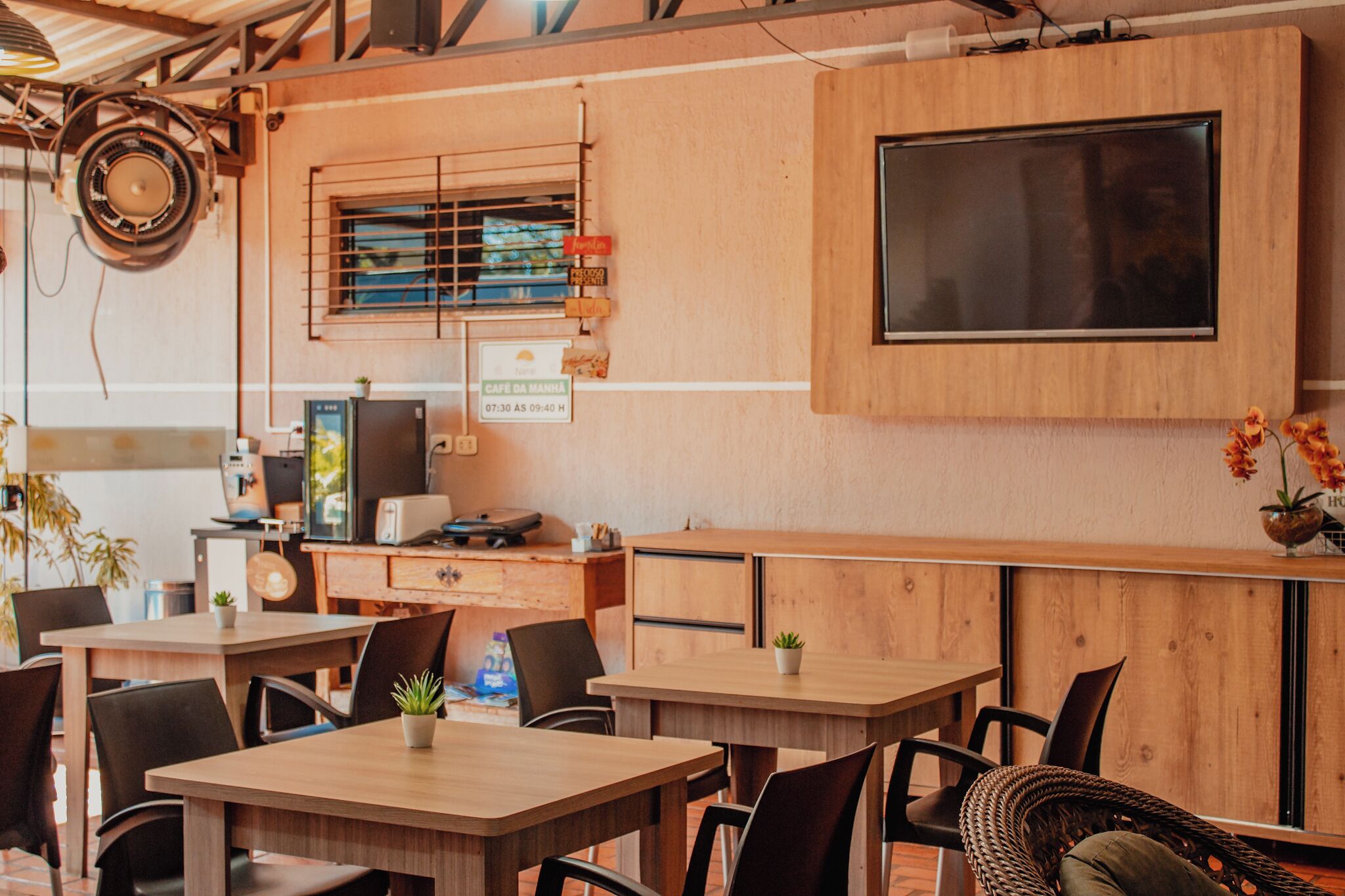 A modern cafe with wooden furniture, a flat-screen TV, and a sign indicating it is open from 07:30 to 09:40.