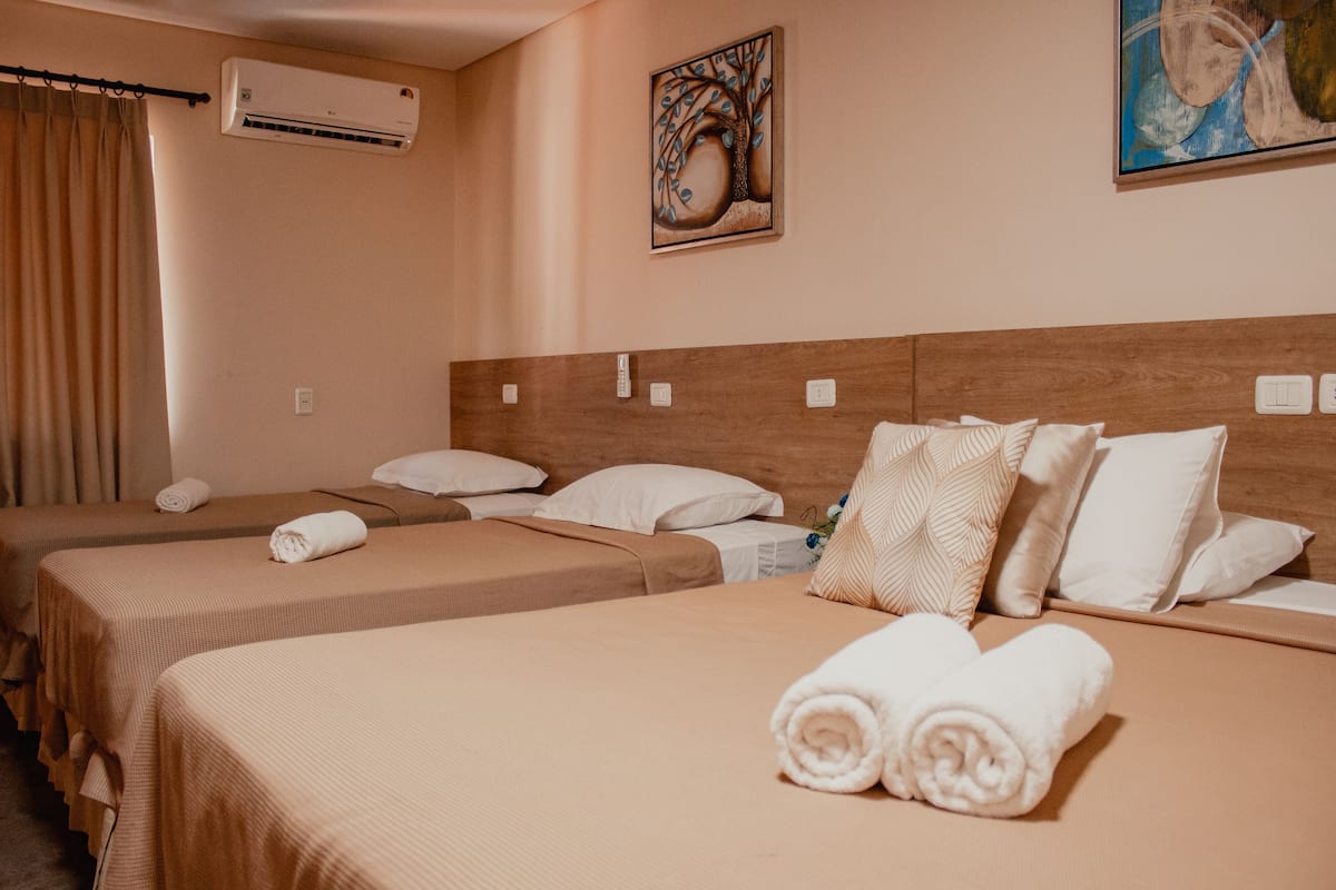 A hotel room with two single beds, each with a brown bedspread and white pillows. There is an air conditioning unit on the wall and a painting above the headboard.