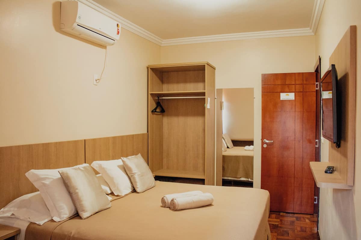 A hotel room with a bed, a wooden wardrobe, a television, and an air conditioning unit.